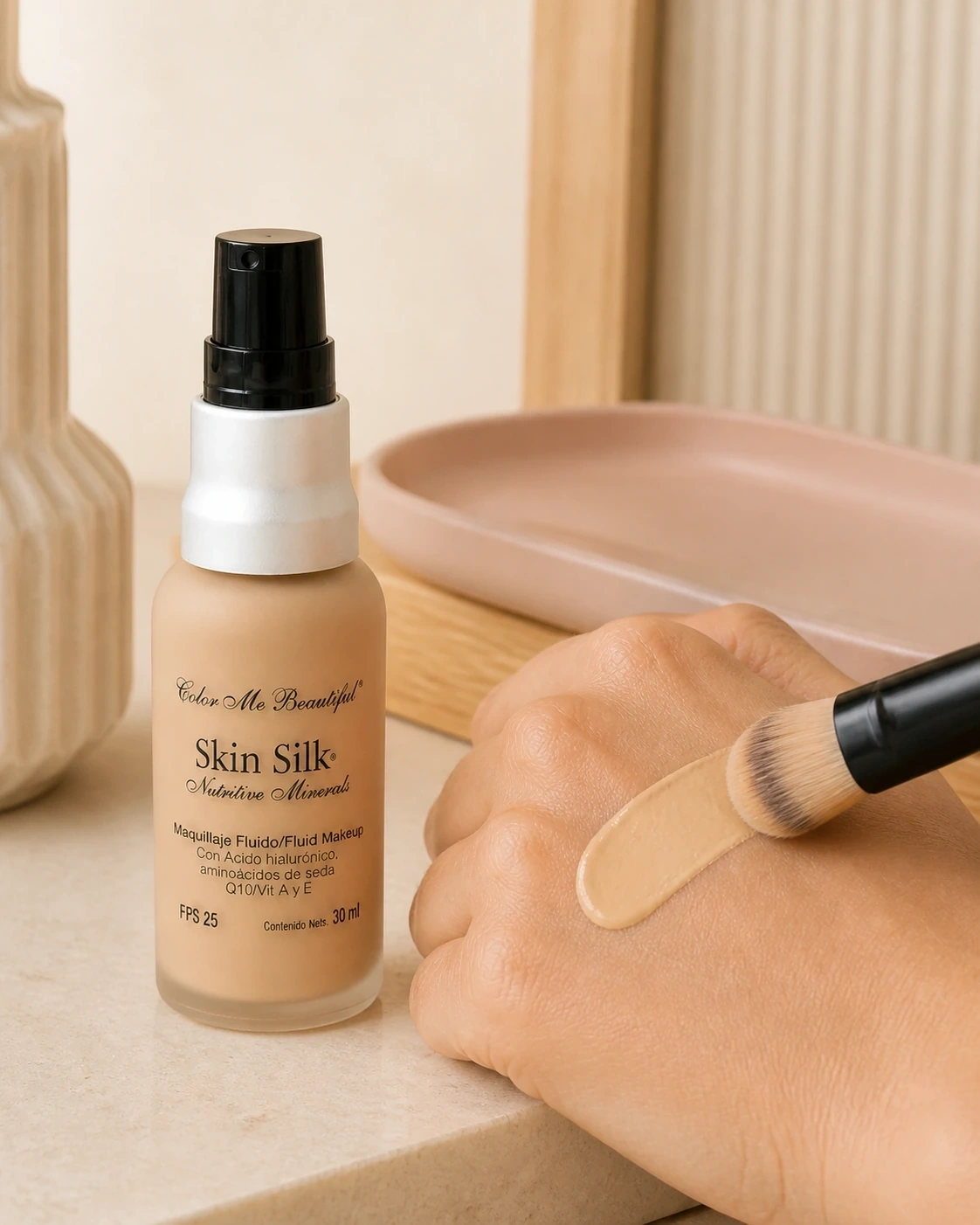 Frasco de maquillaje fluido junto a una muestra beige aplicada con brocha sobre la mano