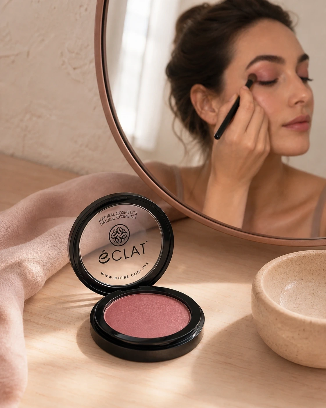 Sombra Eclat en compacto negro sobre tocador con modelo aplicando sombra rosa malva frente a un espejo